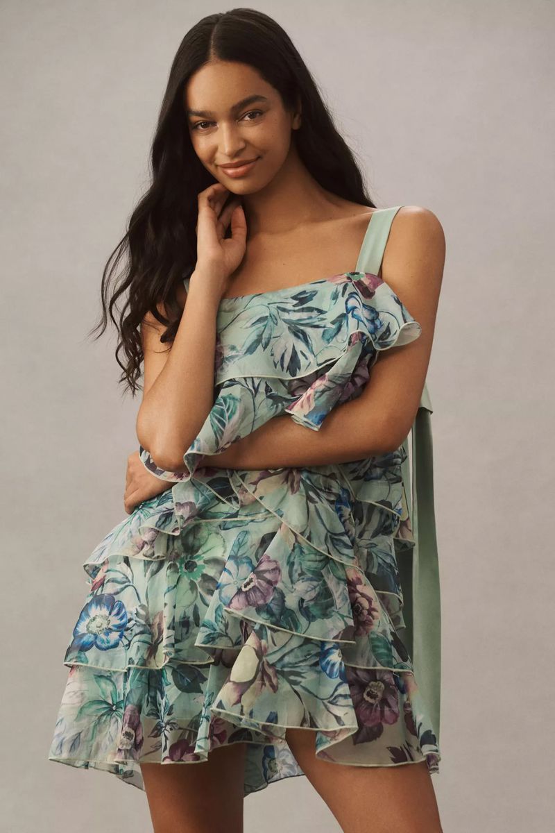 A young, smiling woman poses indoors wearing a floral-patterned, one-shoulder mini dress with ruffle detail against a neutral backdrop.