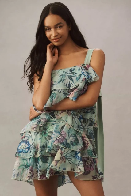 A young, smiling woman poses indoors wearing a floral-patterned, one-shoulder mini dress with ruffle detail against a neutral backdrop.