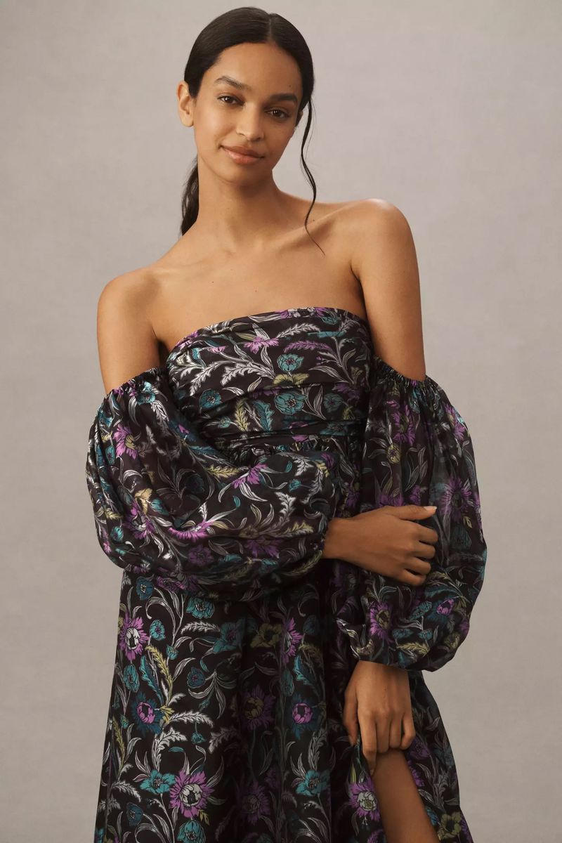 A woman poses in a black off-the-shoulder dress with a floral pattern and puffy sleeves against a neutral background in a studio setting.