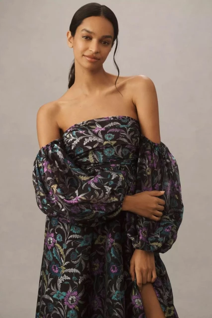 A woman poses in a black off-the-shoulder dress with a floral pattern and puffy sleeves against a neutral background in a studio setting.