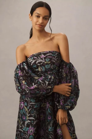 A woman poses in a black off-the-shoulder dress with a floral pattern and puffy sleeves against a neutral background in a studio setting.