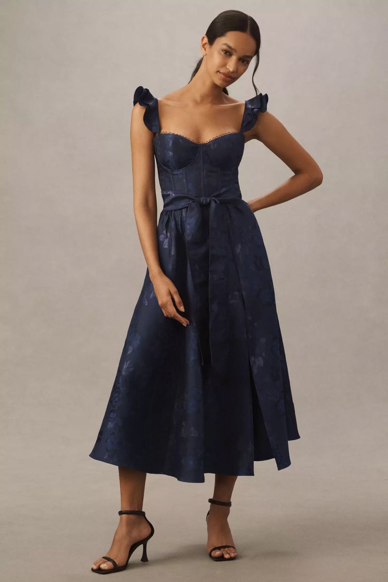 Elegant adult woman in a navy blue floral print dress with shoulder ruffles and strappy heels, posing in a studio. Fashion editorial.