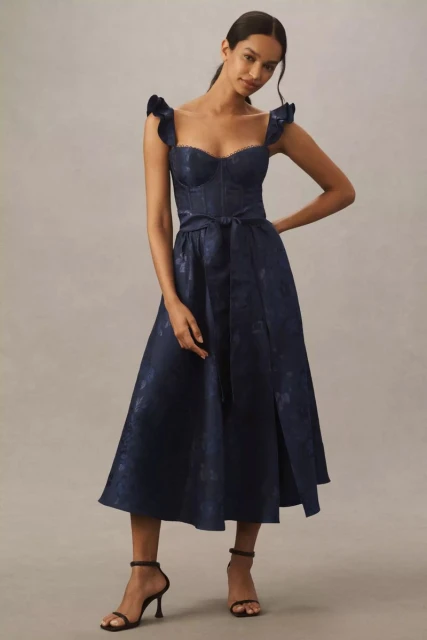Elegant adult woman in a navy blue floral print dress with shoulder ruffles and strappy heels, posing in a studio. Fashion editorial.