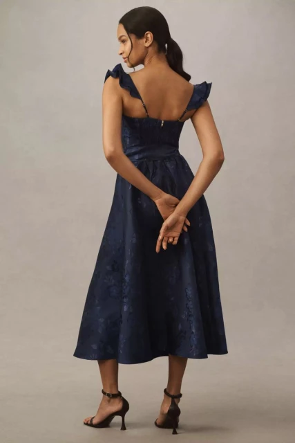 Adult woman in a navy floral midi dress and heeled sandals, photographed from behind in a studio against a neutral backdrop.