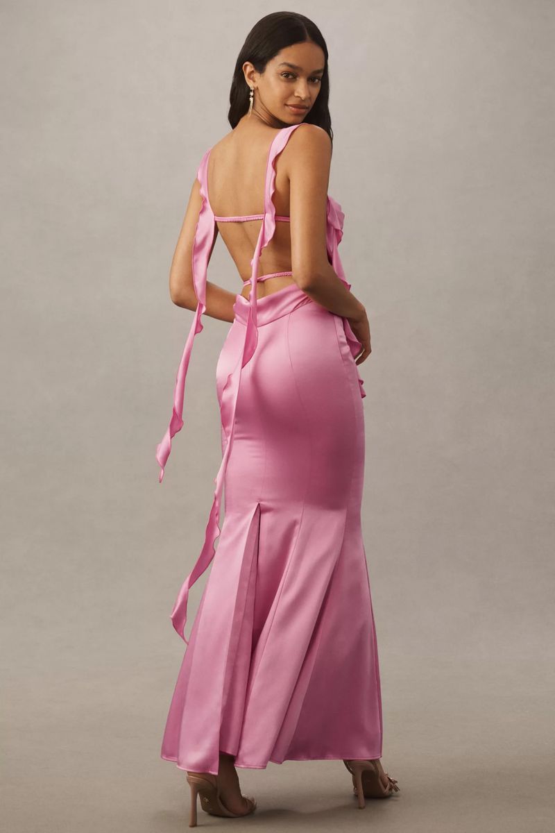 An adult female poses indoors in a pink, floor-length gown with ribbons; she is turned to face the camera with an indoor background.