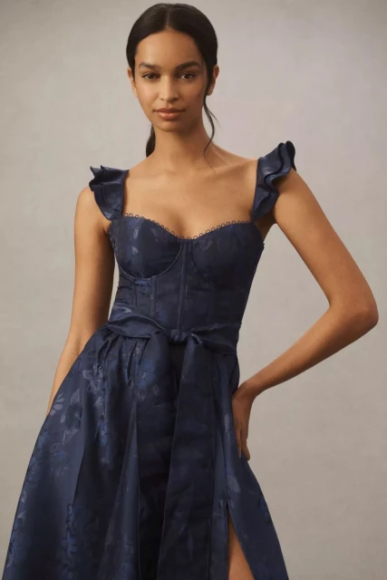 An adult woman poses in a navy blue, floral-patterned dress with ruffles and a belt, set against a neutral studio backdrop.