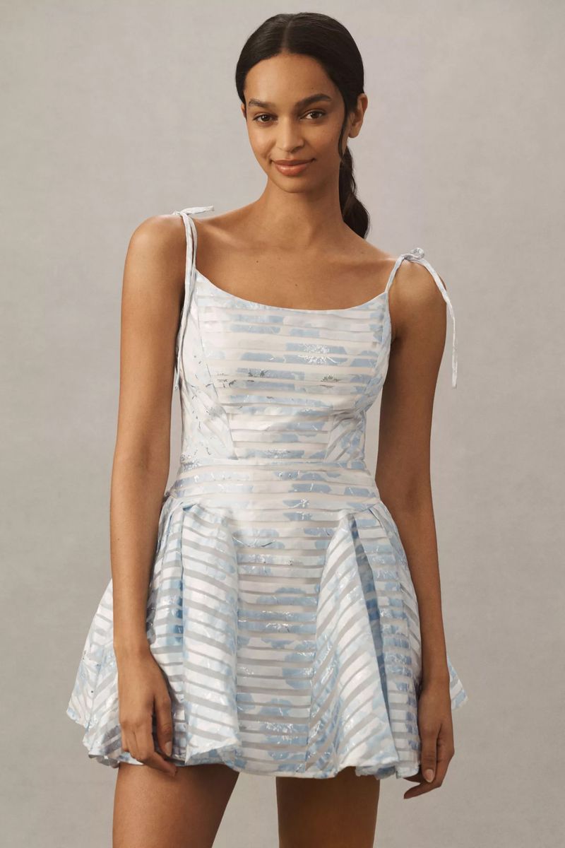A smiling woman poses indoors wearing a light blue and white strapless mini dress with tie shoulder straps against a neutral background.