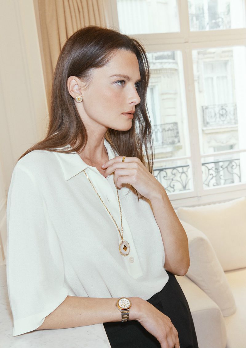Brunette woman in a cream blouse and black skirt wearing gold earrings, necklace, ring and watch poses in a bright room.