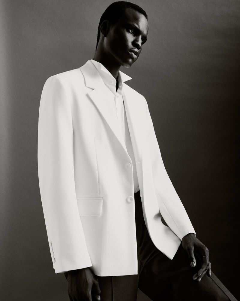 An African male model in a white suit and dress shirt poses against a gray background, showcasing fashion and style.