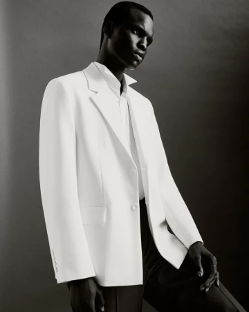 An African male model in a white suit and dress shirt poses against a gray background, showcasing fashion and style.