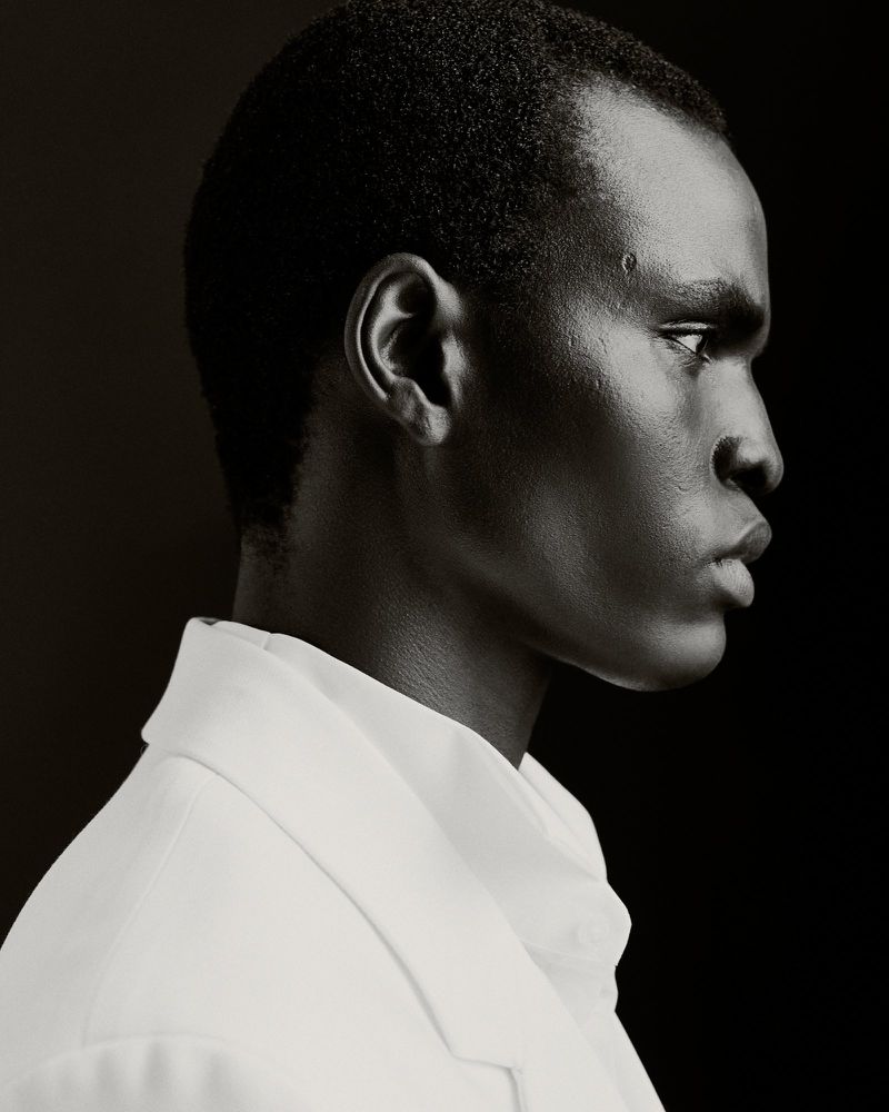 Black and white portrait of an adult male with a clean-shaven head and wearing a collared shirt against a dark, stark background.