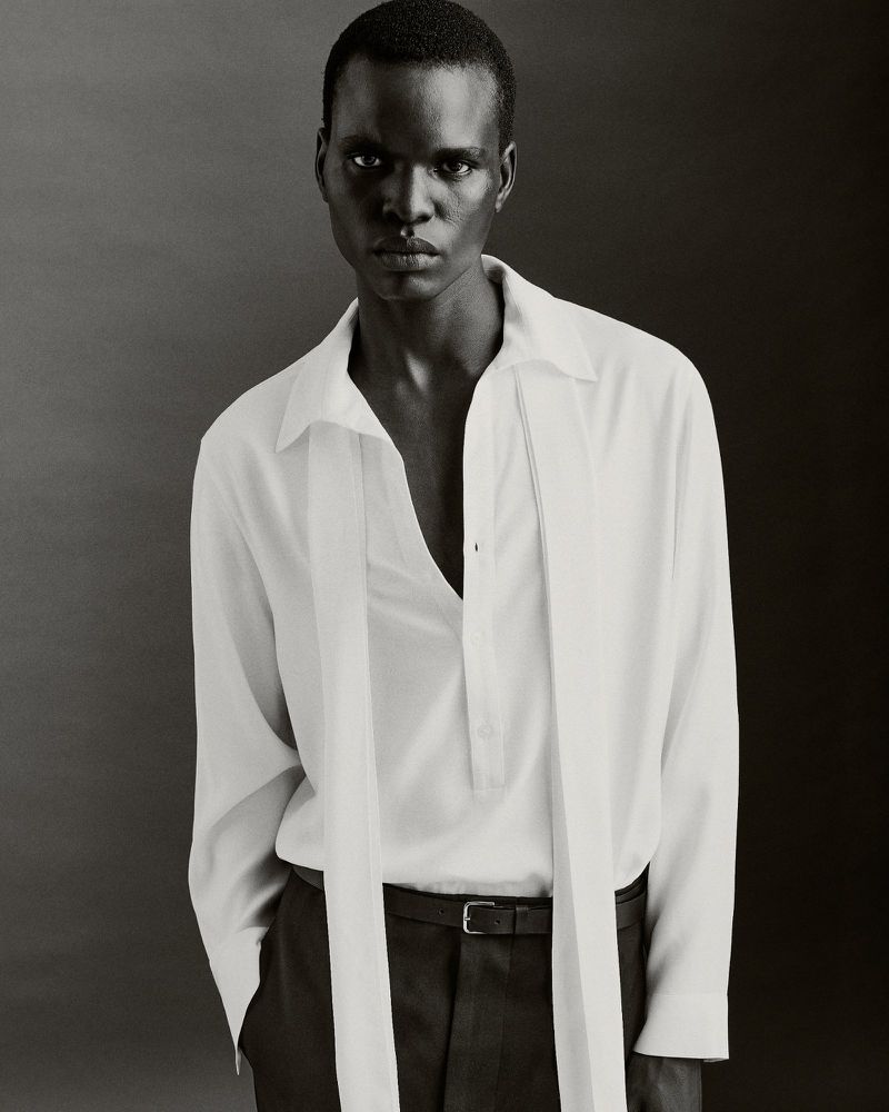 Elegant portrait of a dark-skinned male model in a white collared shirt and black pants against a grey backdrop, showcasing a fashion-forward look.