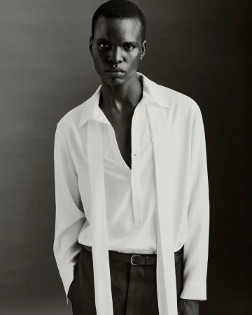 Elegant portrait of a dark-skinned male model in a white collared shirt and black pants against a grey backdrop, showcasing a fashion-forward look.