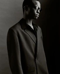 Black man wearing sunglasses and a dark suit jacket against a dark studio background