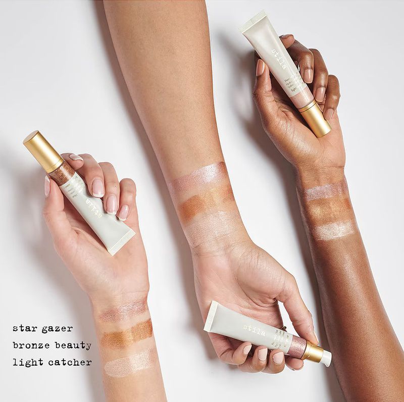 Three women display swatches of liquid eyeshadow on their arms while holding tubes of Stila liquid eyeshadow in an indoor setting.