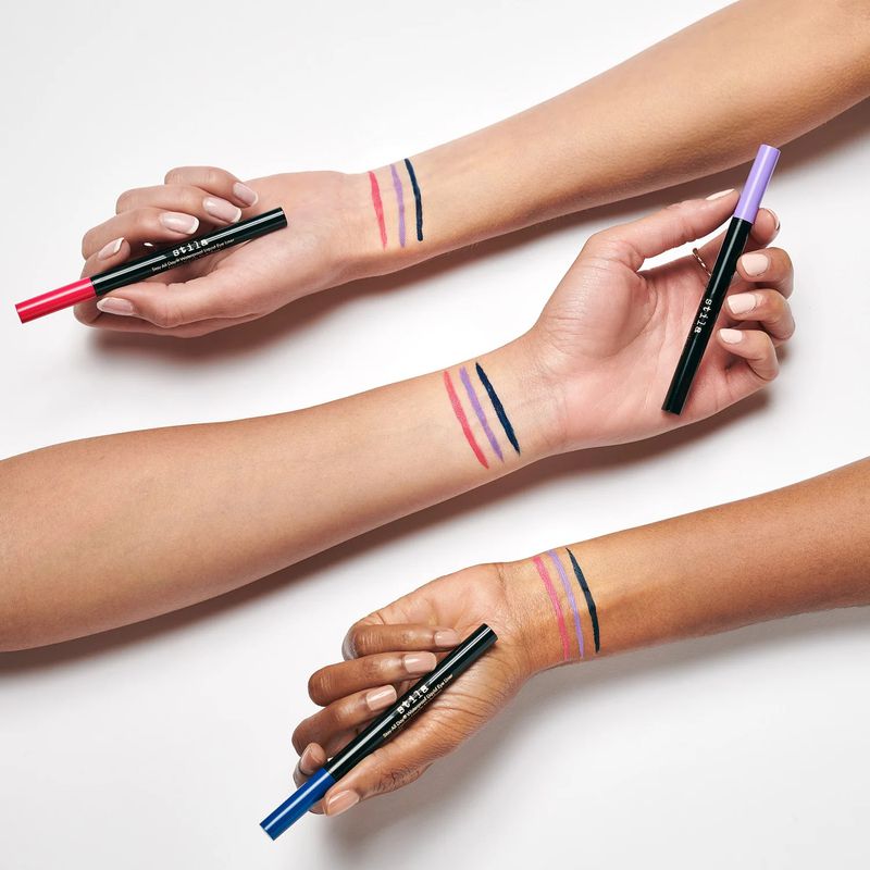 Close-up of three people modeling swatches of colorful eyeliner on their arms, showcasing the beauty product and diverse skin tones.