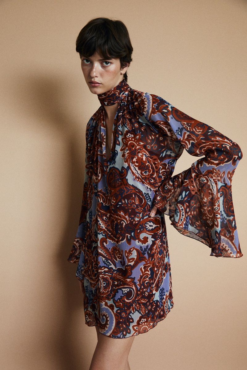 A young woman poses in a studio wearing a brown and blue paisley print dress with long sleeves against a tan backdrop for a fashion editorial.
