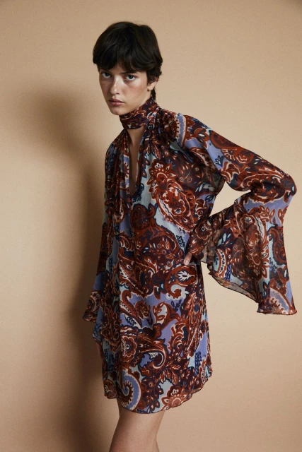 A young woman poses in a studio wearing a brown and blue paisley print dress with long sleeves against a tan backdrop for a fashion editorial.
