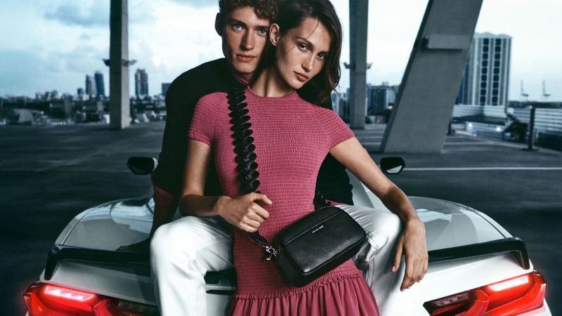 Fashion shot of a stylish couple posing on a white sports car with a city skyline backdrop. The woman wears a pink dress and crossbody bag.