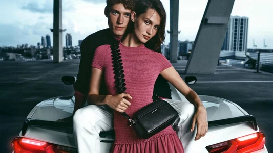 Two models posing by a white car outdoors with an urban skyline in the background, captured for the Michael Kors Resort Campaign.