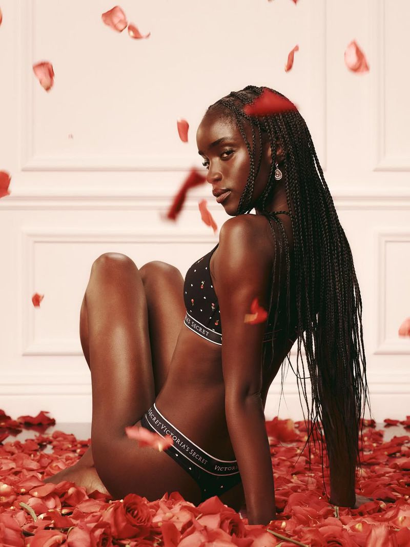 A dark-skinned female model with long braids poses in a bra and panty set, seated among scattered red rose petals against a pale wall.