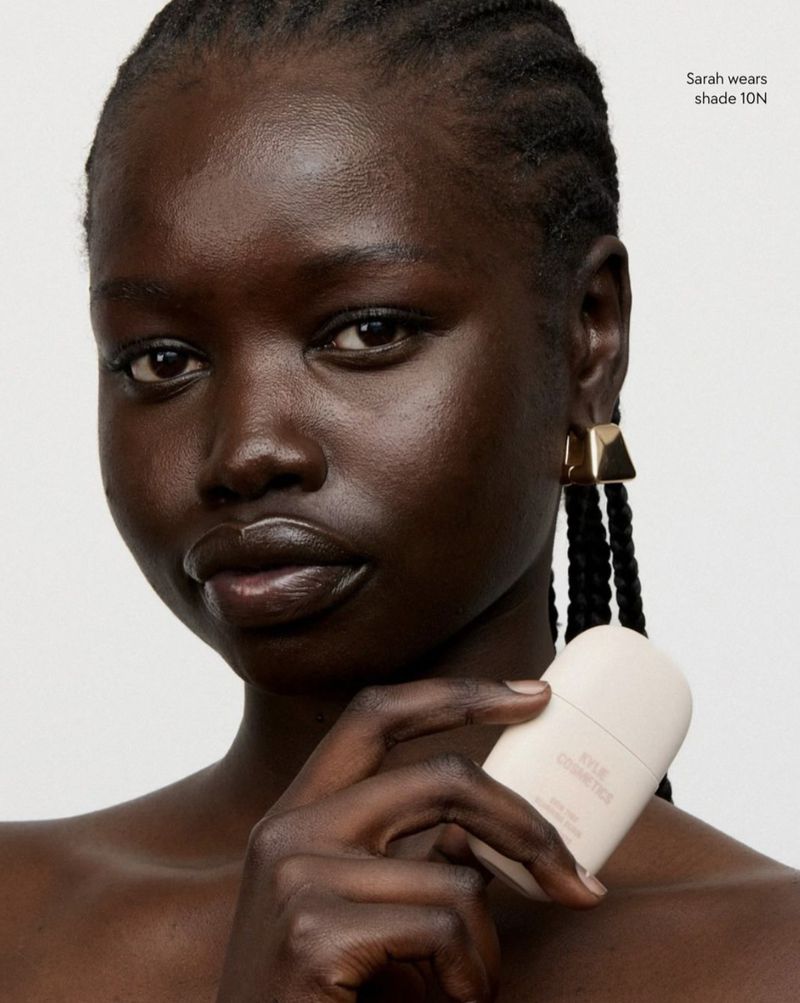 Close-up of a dark-skinned woman with braided hair holding a light-beige cosmetic bottle against a neutral studio backdrop, beauty shot.
