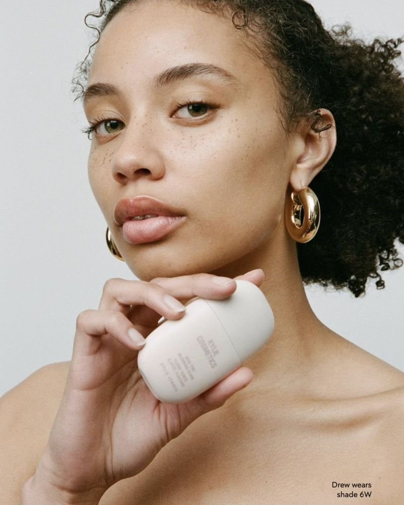 Close-up of a young woman with curly hair, wearing gold hoop earrings, holding a light colored cosmetic bottle against a plain background.