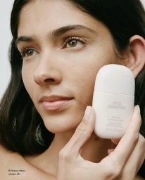Close-up beauty portrait of a woman holding a white Kylie Cosmetics Skin Tint Blurring Elixir bottle near her cheek