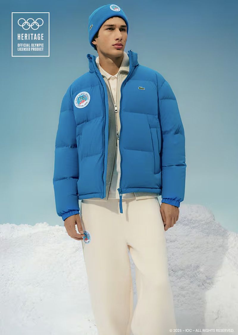 Man poses in blue Lacoste puffer jacket, beanie, and cream sweatpants against a light blue backdrop with snow-covered foreground.