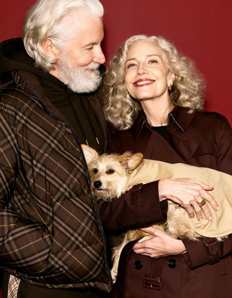 An older couple poses indoors with a small dog wearing a light brown dog coat, with a red background, woman in a brown trench coat.