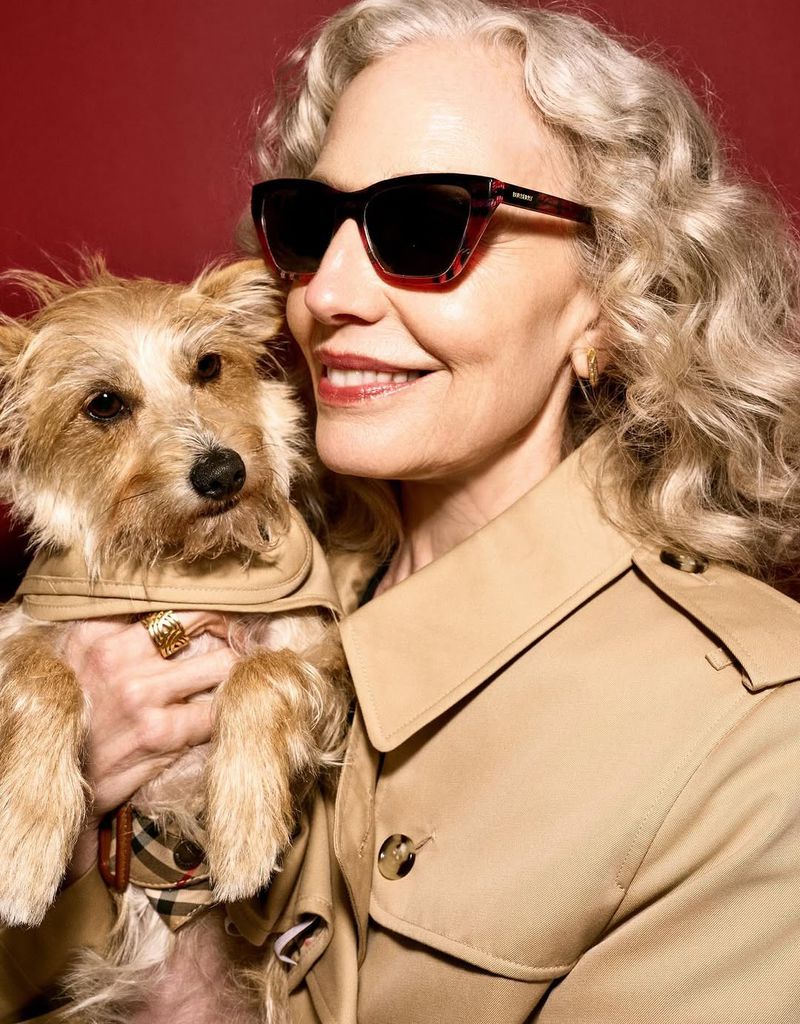 Portrait of an adult female wearing sunglasses and a tan trench coat while holding a small dog also wearing a matching coat in studio.