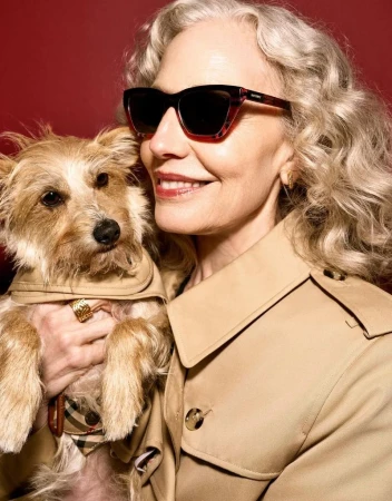 Smiling mature woman with blonde wavy hair wearing sunglasses and a beige trench coat while holding a small dog against a red background.