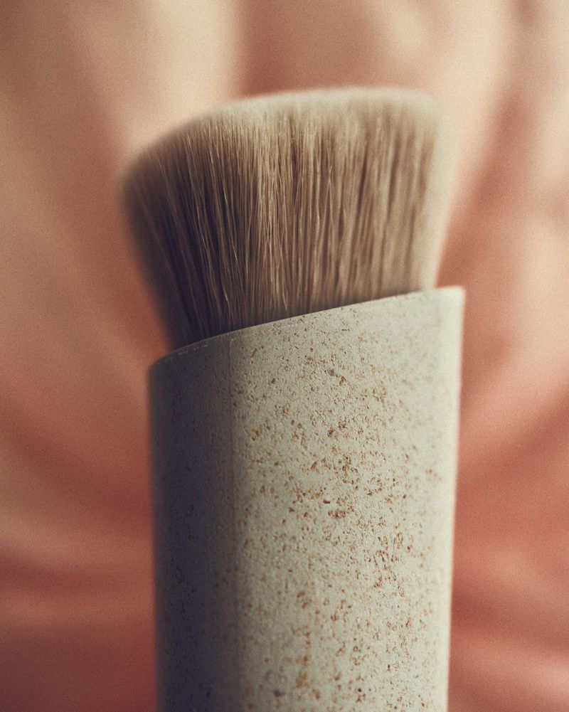 Close-up studio shot of an angled makeup brush with tan bristles and a beige speckled handle against a soft pink background.