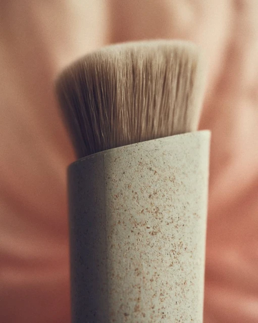 Close-up studio shot of an angled makeup brush with tan bristles and a beige speckled handle against a soft pink background.