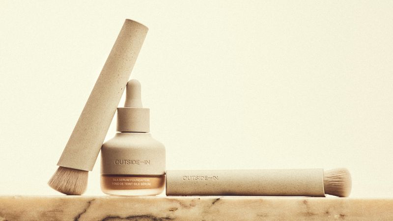 Beauty still life featuring Outside-In brand foundation bottle and two makeup brushes on a marble surface against a cream background.