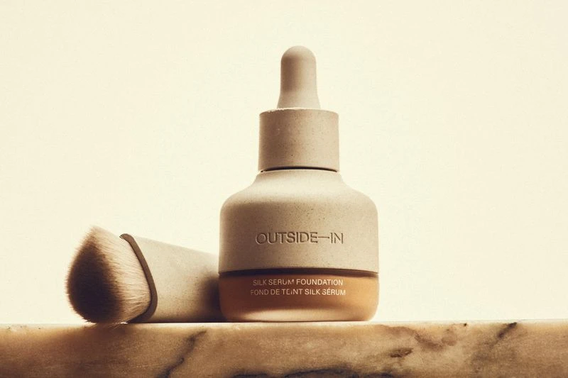 Cosmetic product shot: neutral-toned foundation bottle with dropper and applicator brush on a marble surface, against a light background.