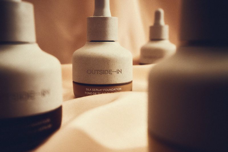 Three bottles of Outside-In Silk Serum Foundation are displayed on a soft peach-colored fabric, arranged for a beauty product shot.