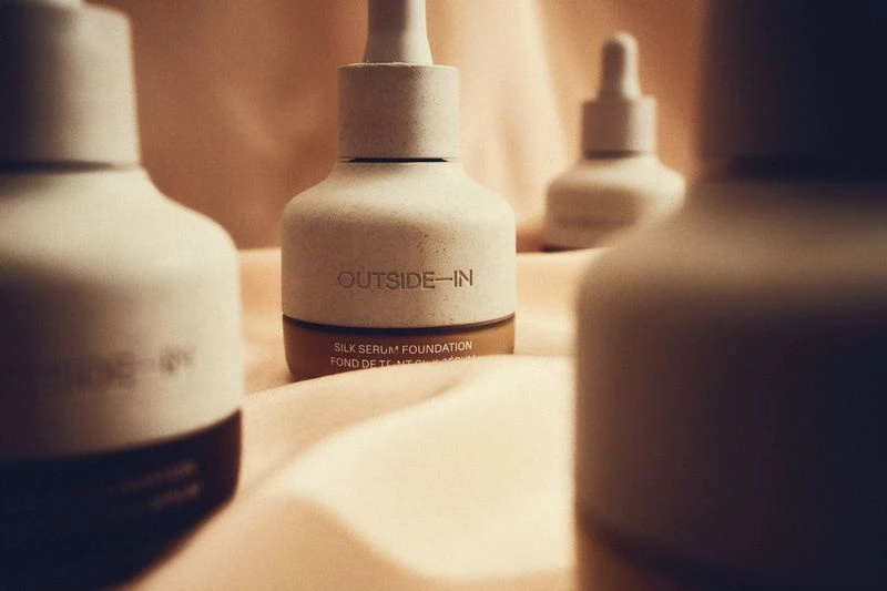 Three bottles of Outside-In Silk Serum Foundation are displayed on a soft peach-colored fabric, arranged for a beauty product shot.
