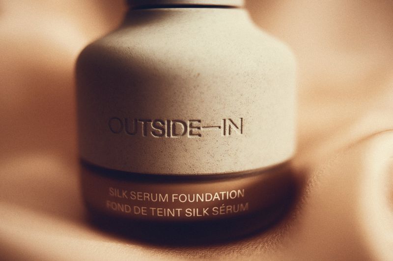 Close-up of a bottle of Outside-In Silk Serum Foundation against a draped fabric backdrop, showcasing its neutral beige tones and texture.