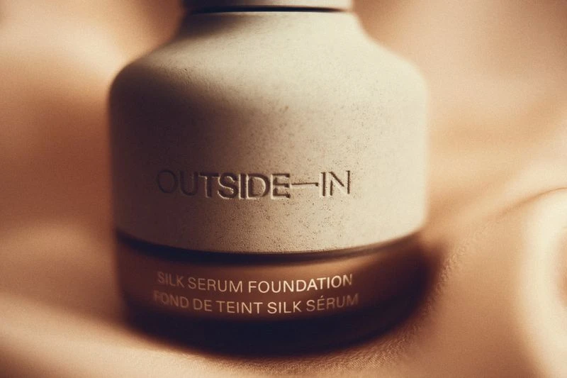 Close-up of a bottle of Outside-In Silk Serum Foundation against a draped fabric backdrop, showcasing its neutral beige tones and texture.