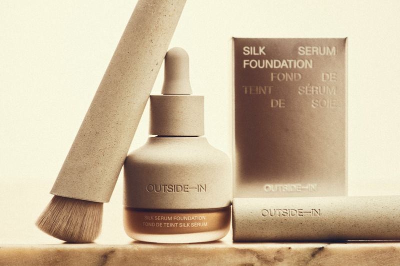 Neutral studio shot of Outside-In brand silk serum foundation bottle, box, and brush with sand texture, soft light, and neutral color palette.