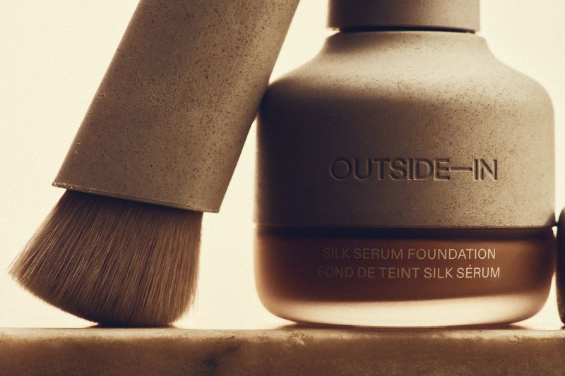 Close-up shot of a foundation bottle and makeup brush. The label 'OUTSIDE-IN' is visible on the bottle, in neutral color palette.