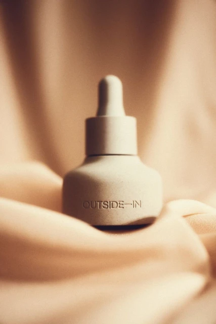 Close-up of a grey skincare serum bottle on a draped tan satin background in diffused lighting, featuring the text 'OUTSIDE-IN'.