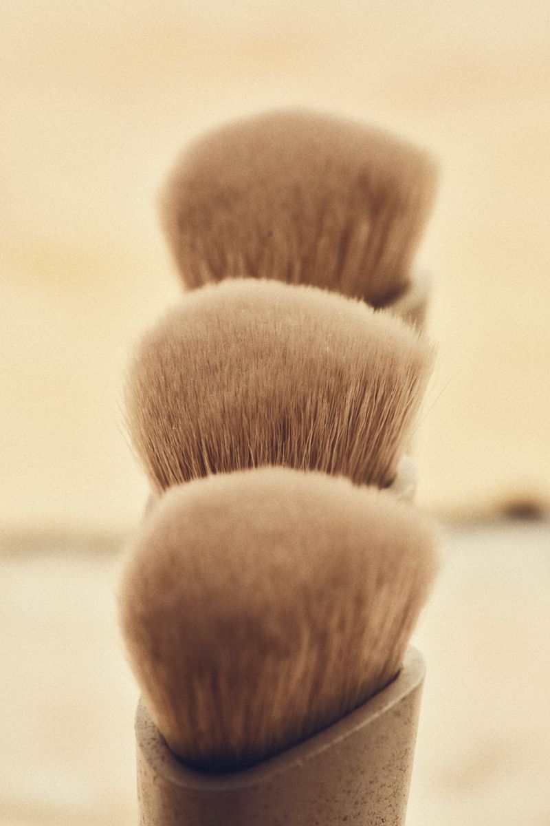 Close-up of three beige makeup brushes stacked vertically, soft focus, against a light background.