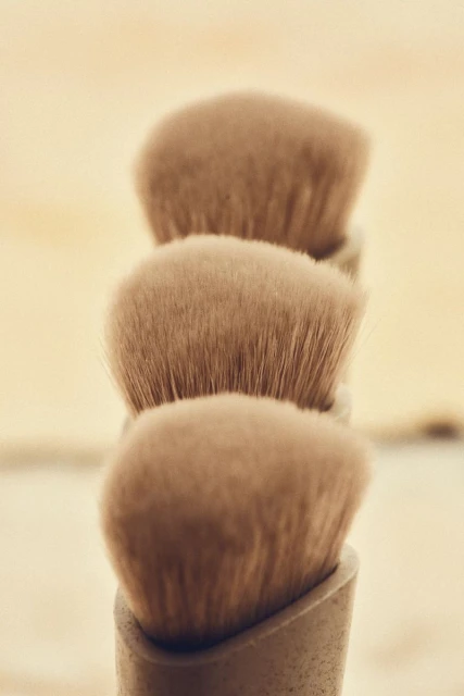 Close-up of three beige makeup brushes stacked vertically, soft focus, against a light background.