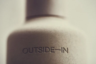 Close-up of a matte beige textured cosmetic bottle featuring debossed Outside In typography in soft ambient light.