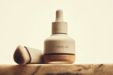 A bottle of Outside In Silk Serum Foundation with a matching angled makeup brush on a marble surface against a warm cream backdrop.