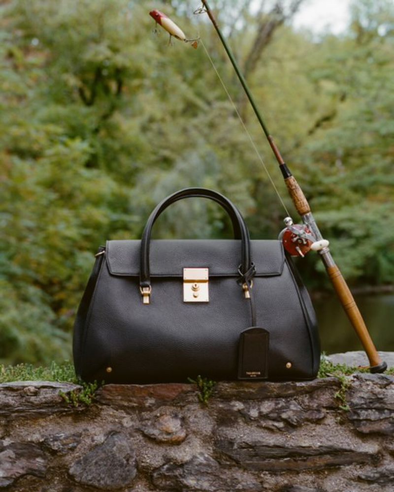 Black leather handbag with gold hardware and a fishing rod backdrop against a blurred green forest background.