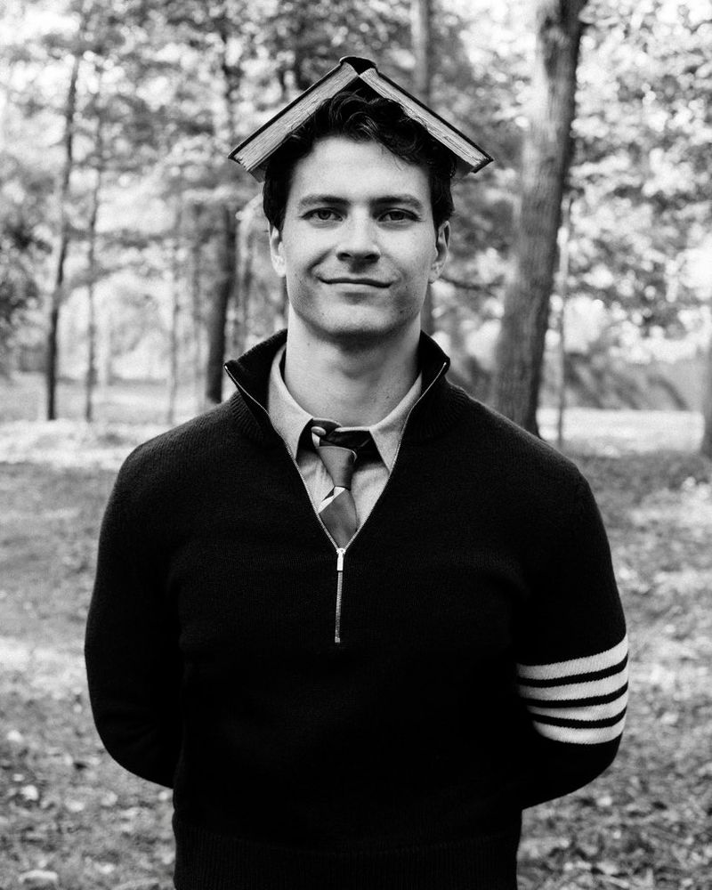 Black and white portrait of a man in a zip-up sweater balancing an open book on his head outdoors in a forest.