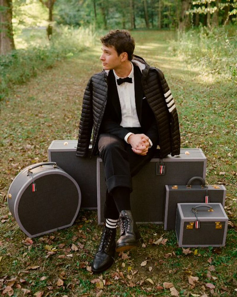 Young man in a suit and puffer jacket poses outdoors seated on a stack of vintage suitcases in a wooded setting.